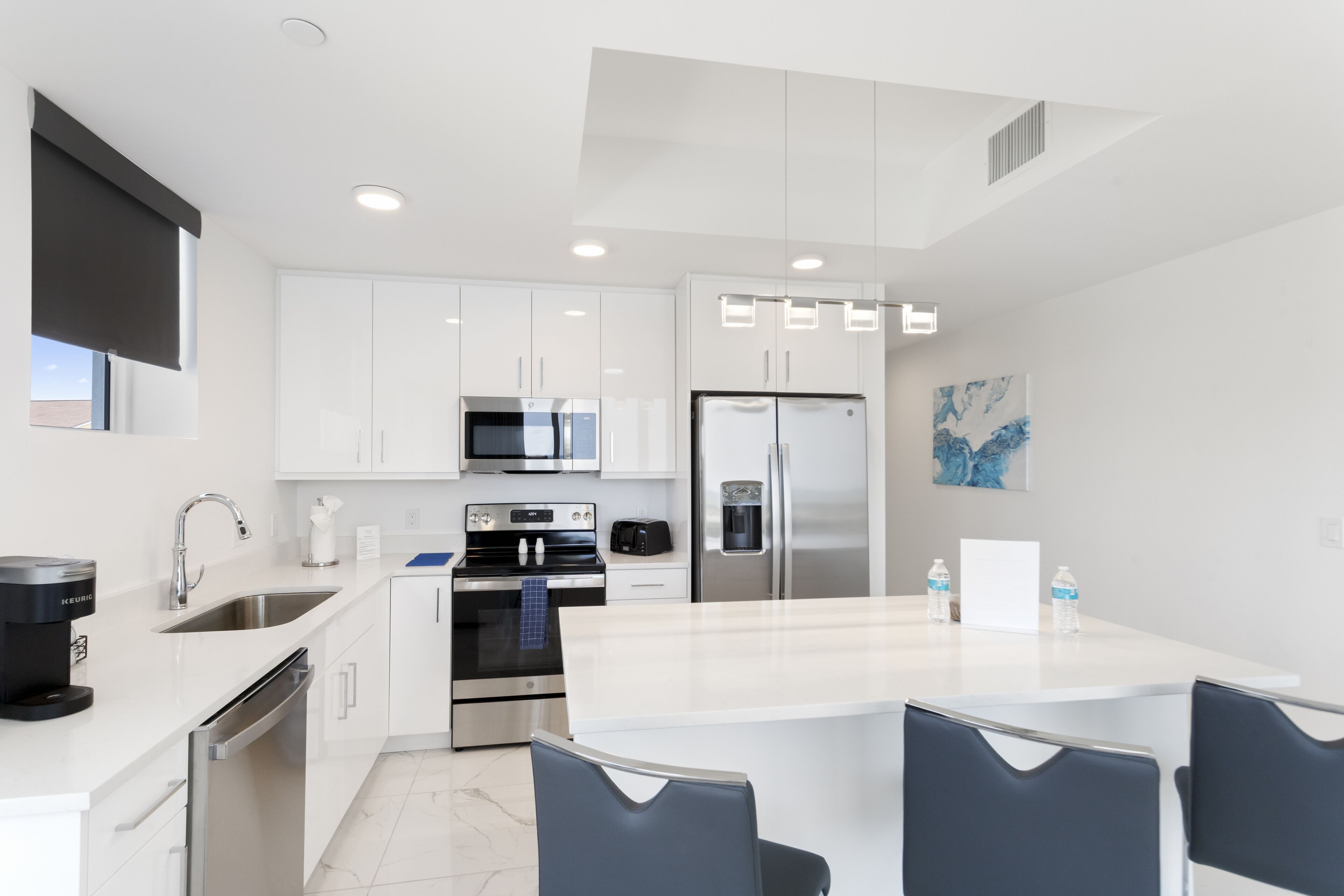 Una cocina moderna con muebles blancos, electrodomésticos de acero inoxidable, una isla central con taburetes y un amplio ventanal.