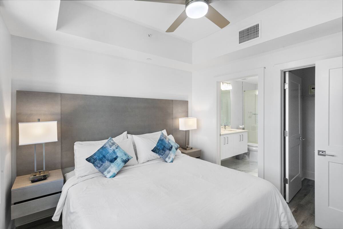 A modern hotel room with a large bed, two bedside tables with lamps, a ceiling fan, and a bathroom visible through an open door.