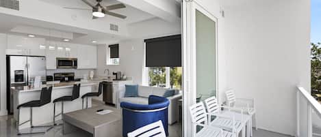 A modern kitchen with white cabinets, a microwave, and a coffee machine. There is a dining area with white chairs and a blue sofa.