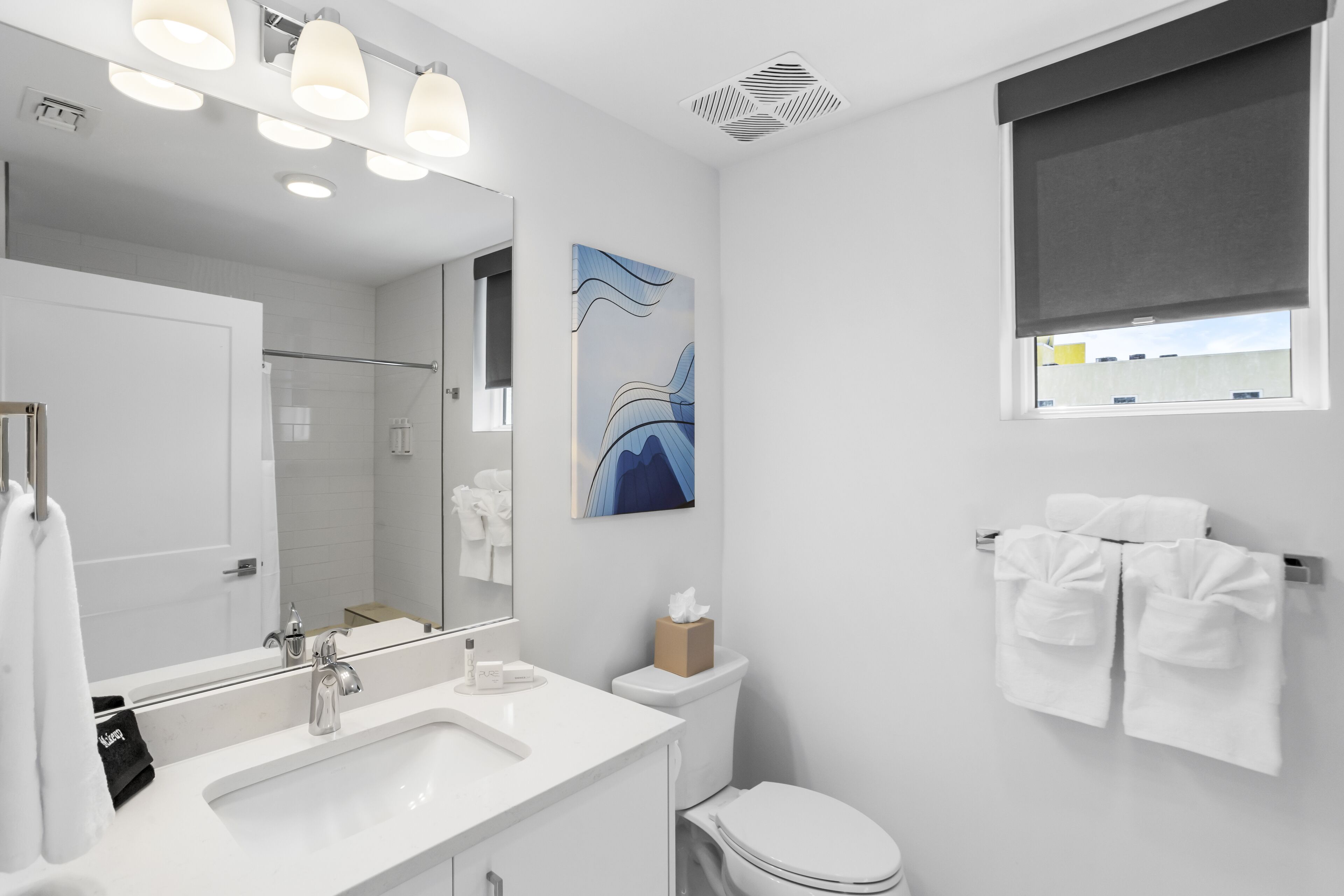 A modern bathroom with a white sink, mirror, and toilet. There is a towel rack with towels and a painting on the wall.