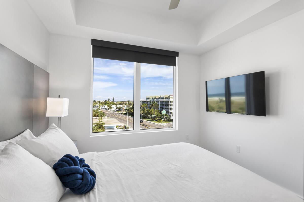 A modern hotel room with a large bed, a flat-screen TV mounted on the wall, and a window offering a city view.