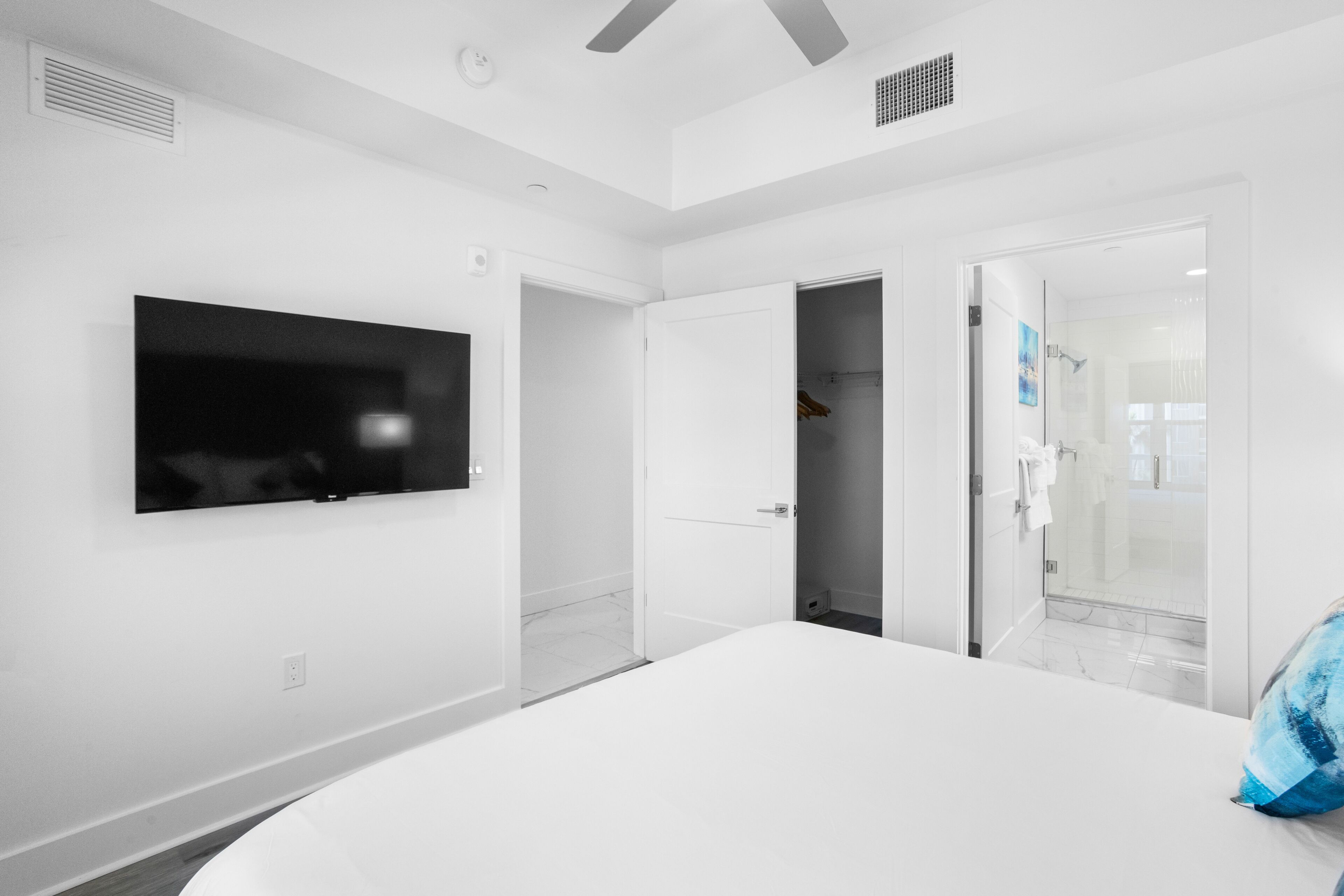 A modern hotel room with a large bed, a flat-screen TV mounted on the wall, a walk-in closet, and a glass shower enclosure.