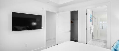 A modern hotel room with a large bed, a flat-screen TV mounted on the wall, a walk-in closet, and a glass shower enclosure.