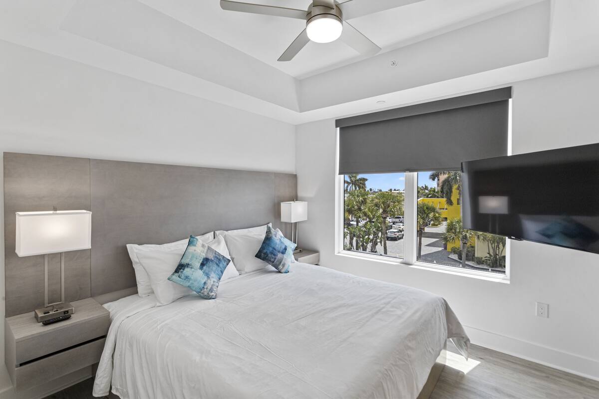 A modern hotel room with a large bed, a nightstand with a lamp, a TV mounted on the wall, and a window with a view of the outside.
