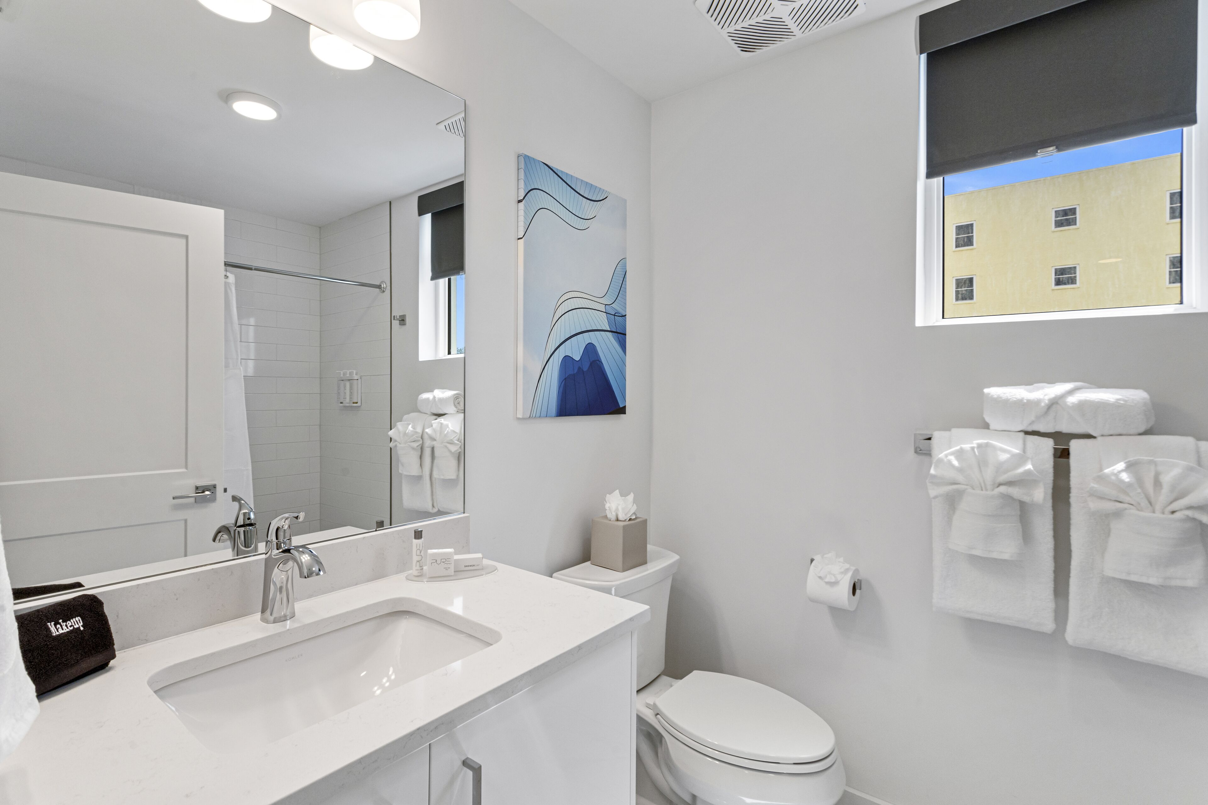 Un baño moderno con un lavamanos blanco, un espejo, un inodoro y una ventana con vista a un edificio.