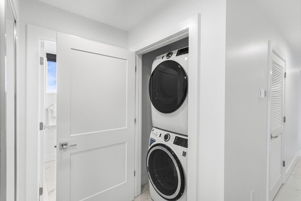 Un cuarto de lavandería con dos lavadoras y secadoras apiladas, paredes blancas y una puerta que da a otra habitación.