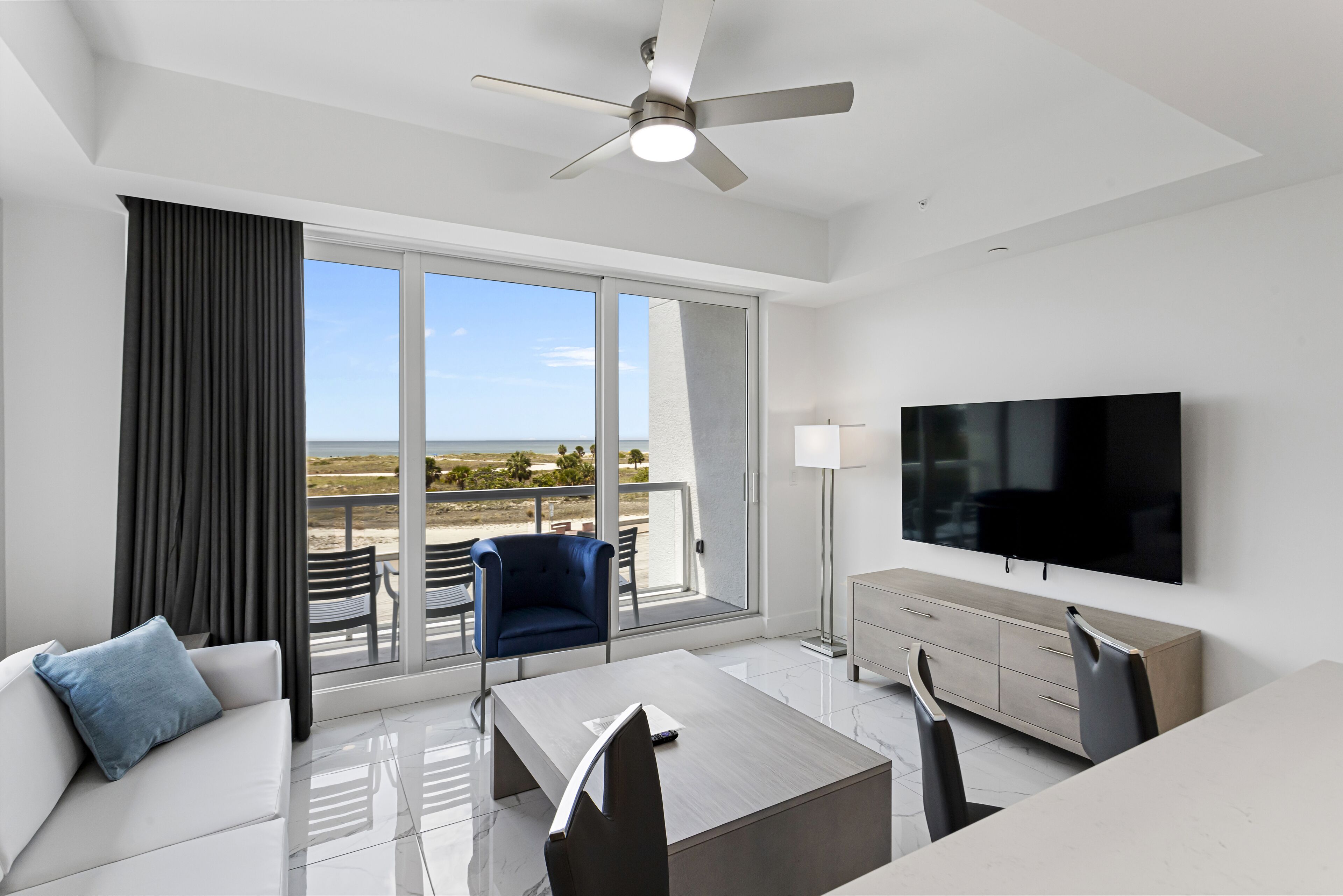Een moderne hotelkamer met balkon, flatscreen-tv, bank, eettafel en plafondventilator.
