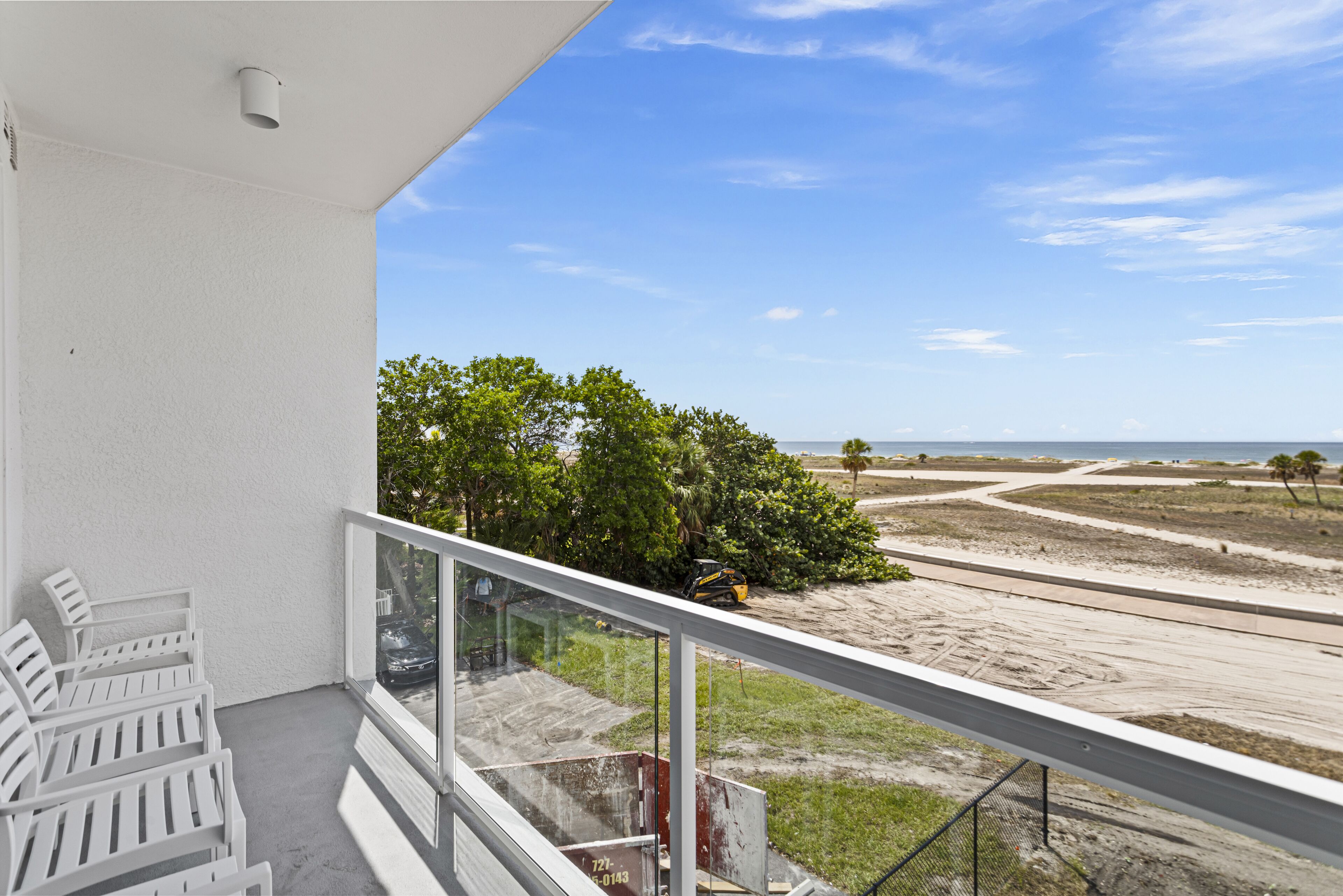 A balcony with white chairs, a glass railing, and a view of a beach with trees and a clear sky.