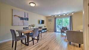A modern living room with a dining area, a flat-screen TV mounted on the wall, and a large window offering a view of the outdoors.