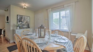 Un coin repas avec une table ronde dressée pour quatre personnes, des chaises en bois, une nappe blanche et une fenêtre avec des stores.