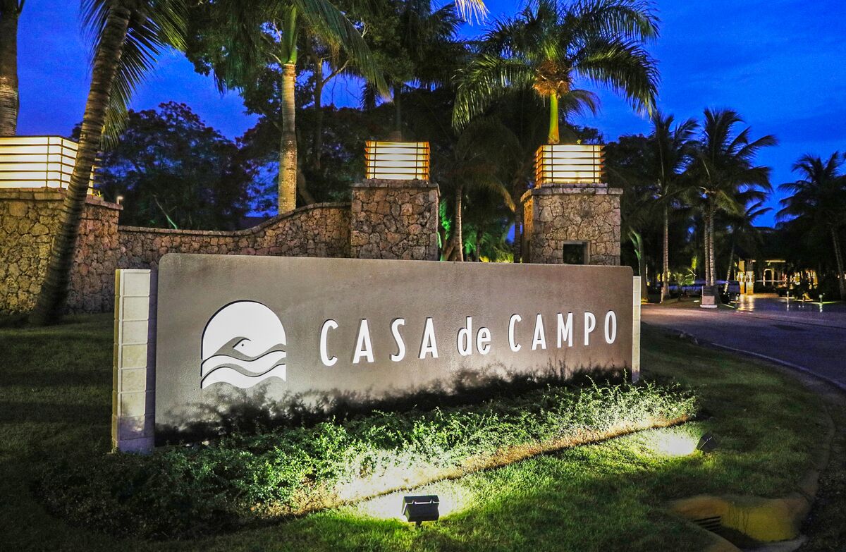 A sign for CASA de CAMPO with a wave logo, surrounded by palm trees and stone walls, indicating a tropical setting.