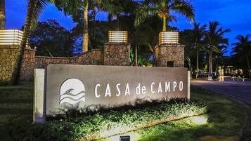 A sign for CASA de CAMPO with a wave logo, surrounded by palm trees and stone walls, indicating a tropical setting.