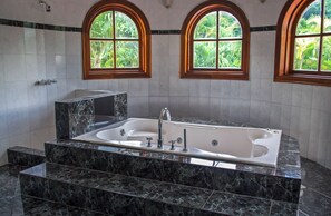 Une salle de bain dotée d’une baignoire en marbre, d’une douche et de deux fenêtres en arc de cercle offrant une vue sur la verdure.