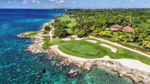 Vista aérea de un campo de golf costero con aguas turquesas cristalinas, una playa de arena y abundante vegetación.