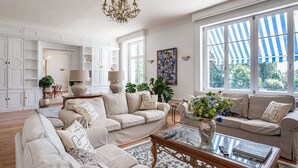 Un salon spacieux, doté d’un grand canapé, d’une table basse et d’un lustre. De grandes fenêtres sont agrémentées d’un auvent à rayures, et un tableau orne le mur.