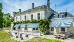 Une grande maison à deux étages, dotée d’une façade en pierre, d’un patio couvert avec une table et des chaises, et d’un petit pont menant à l’entrée.
