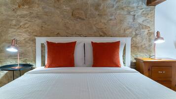Un lit avec une literie blanche et deux oreillers rouges, flanqué de deux lampes de chevet, le tout placé devant un mur en pierre.