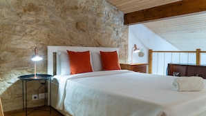A bedroom with a stone wall, a wooden ceiling, a bed with white bedding and orange pillows, a bedside table with a lamp, and a view of the outdoors. - Casa São José (Carvalhal)