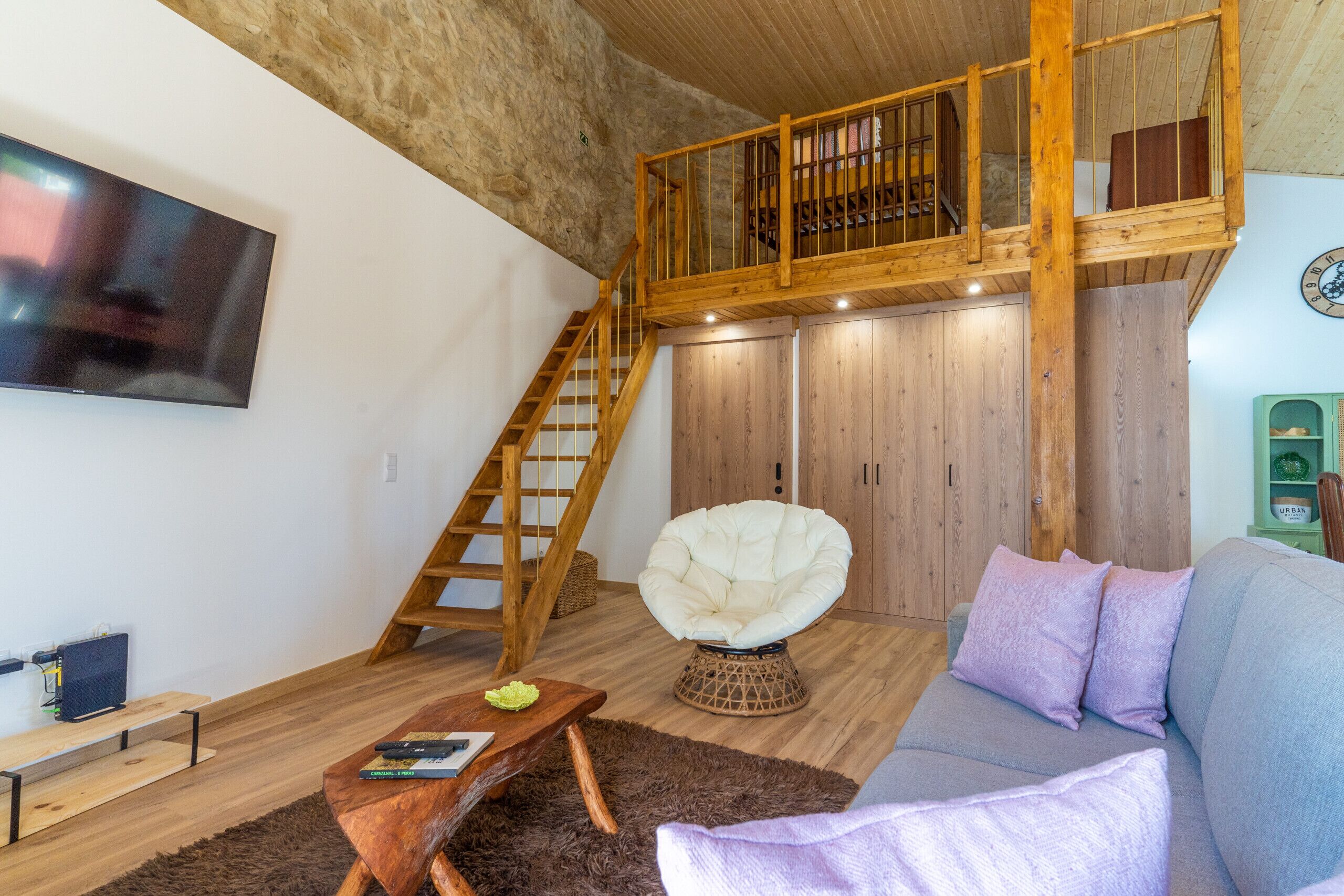 A loft-style living space with a wooden staircase, a white armchair, a sofa with cushions, and a flat-screen TV mounted on the wall.