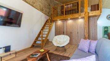 A loft-style living space with a wooden staircase, a white armchair, a sofa with cushions, and a flat-screen TV mounted on the wall.
