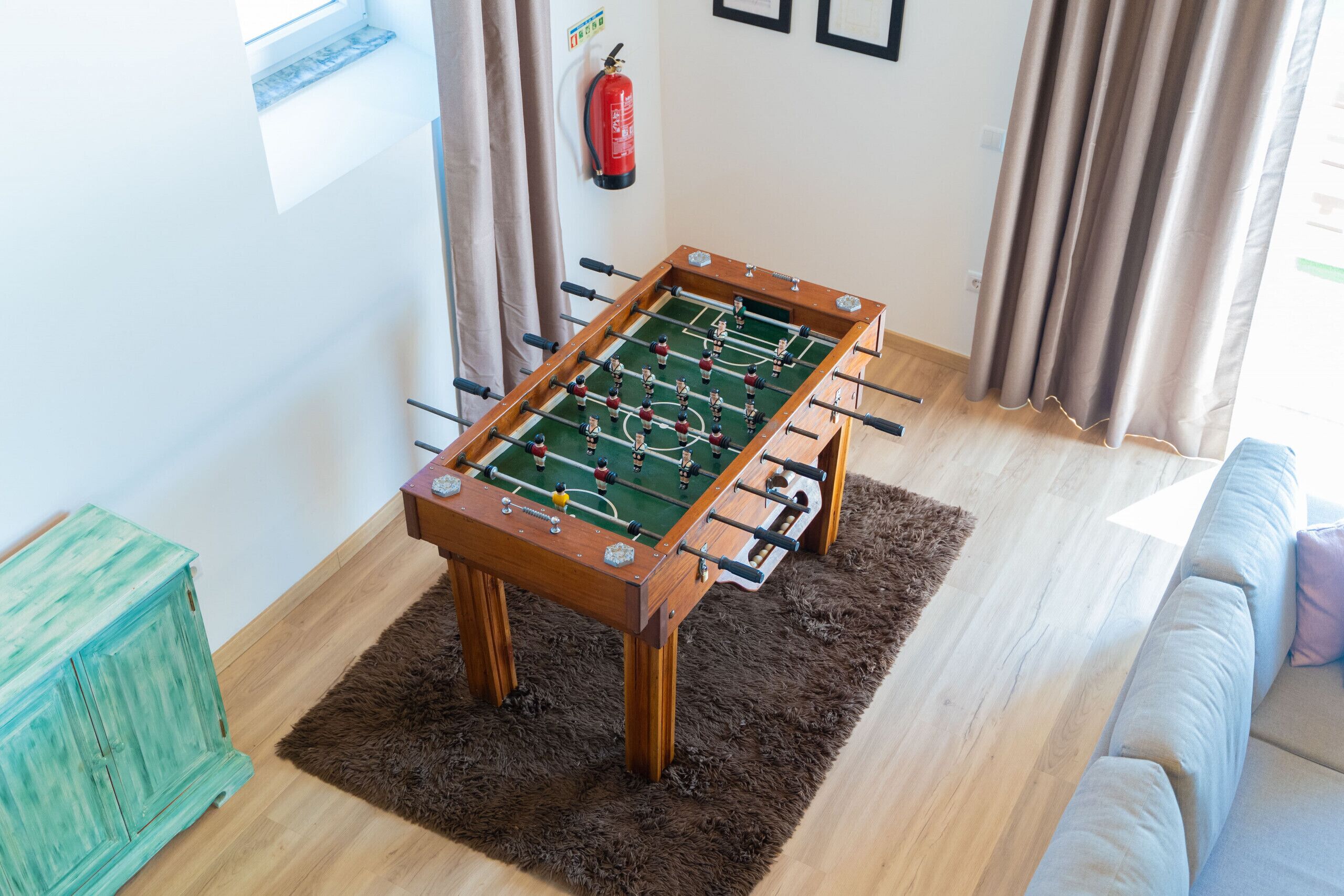 Uma mesa de futebol de bolso em um quarto com piso de madeira, um tapete macio, um armário verde e um extintor de incêndio na parede.