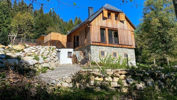 Une cabane en bois, posée sur des fondations en pierre, entourée d’arbres et surplombée par un ciel bleu azur.