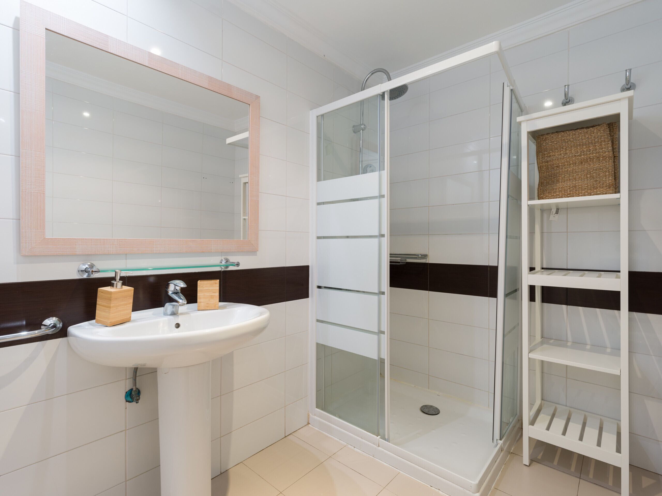 Un baño con un lavamanos blanco, un espejo, una ducha con cerramiento de vidrio y un estante blanco con cestas.