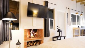 A modern hotel room with a brick fireplace, a flat-screen TV mounted on the wall, a bed with a white comforter, a small table with a lamp, and a window with curtains. - Cabaña El Encino III en Real del Monte (Mineral del Monte)