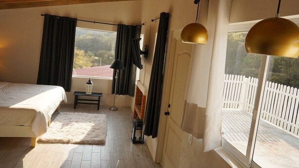 A bedroom with a bed, a small table with a lamp, a window with curtains, and a view of a white fence outside. - Cabaña El Encino III en Real del Monte (Mineral del Monte)