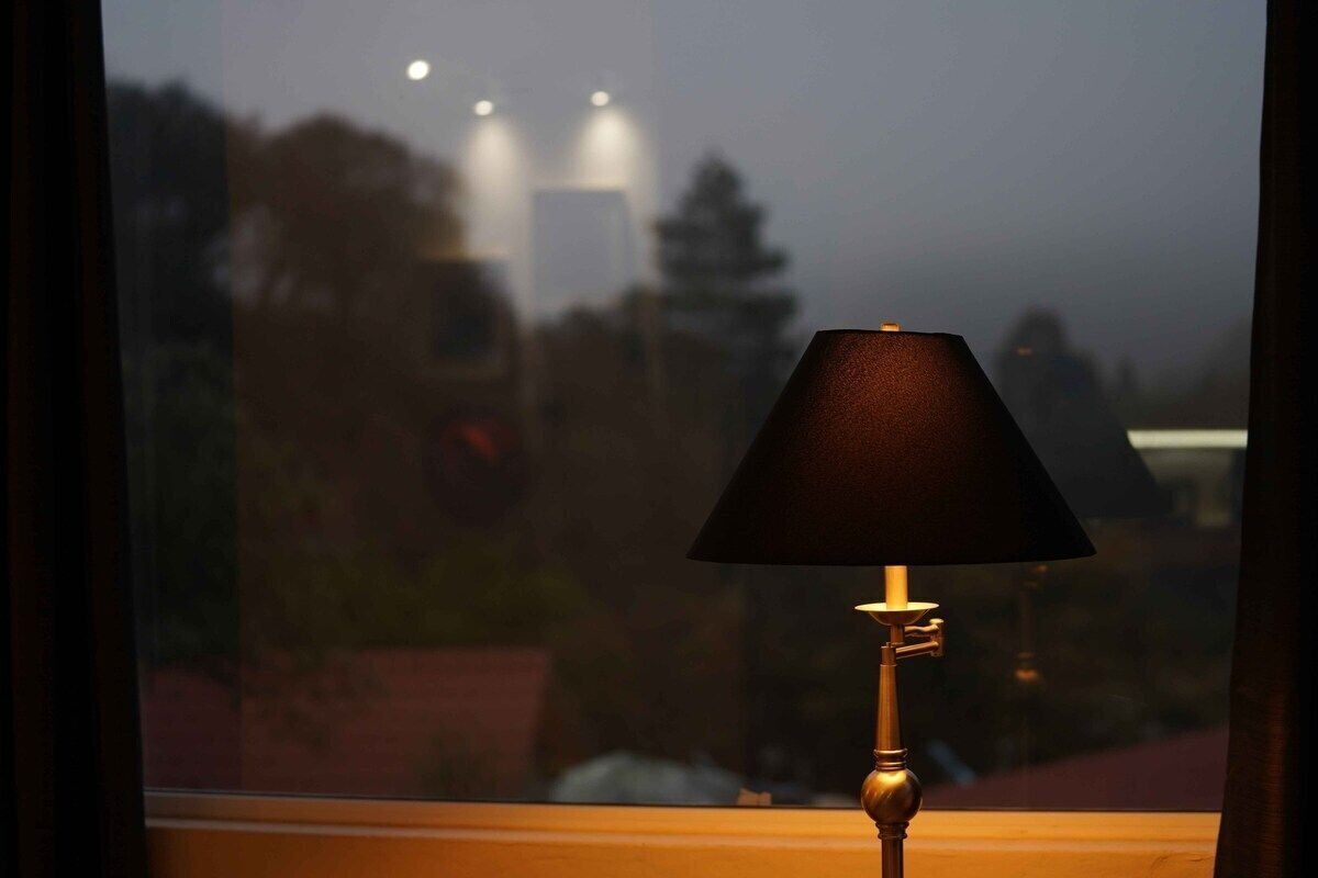 Pöydällä, ikkunan lähellä, on lamppu, ja ikkunasta avautuu näkymä kaupungin siluettiin auringonlaskun aikaan.