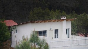 A white building with a red roof, surrounded by a white picket fence and dense forest. - Cabaña El Encino III en Real del Monte (Mineral del Monte)