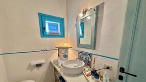 A bathroom with a toilet, sink, and mirror. There is a window above the sink and a door to the right. - Villa Calisto - San Pantaleo (Olbia)