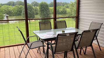 En overdækket terrasse med et bord og stole, med udsigt over en græsplæne og et vandareal.