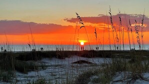Un coucher de soleil sur une plage, avec des silhouettes de personnes et des herbes hautes.