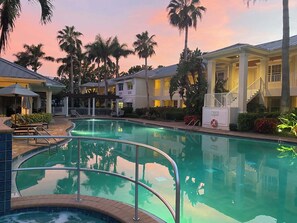 Ein Pool mit geschwungenem Design, umgeben von Palmen und einem Gebäude mit überdachter Terrasse.