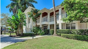 Un immeuble à plusieurs étages, doté d’une façade blanche, de balcons et d’une pelouse bien entretenue agrémentée de plantes tropicales.