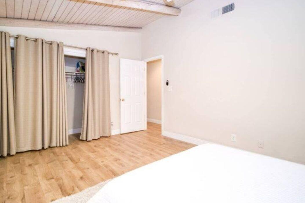 A room with wooden flooring, a bed, beige curtains, and a white door.