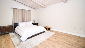 Ein Schlafzimmer mit einem Bett, einem Nachttisch, Holzboden, Deckenventilator und einem Fenster mit Vorhängen.