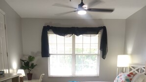 Une chambre à coucher comprenant un lit, un bureau, une chaise, un ventilateur de plafond, une fenêtre avec des rideaux, une lampe et une petite table sur laquelle est posée une plante.