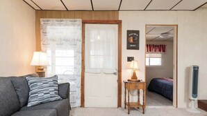 A room with a grey sofa, a wooden side table with a lamp, a door with a nautical-themed curtain, and a bed with a red and white striped blanket. - Lovely- 3 Bedroom apt in Vaca Beach home (Hampton)