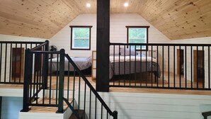 Ein Schlafzimmer im Loft-Stil mit Holzbalkendecke, zwei Fenstern, einem Bett und einer Treppe mit schwarzen Geländern.
