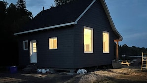Ein niedriges, dunkel gefärbtes Haus mit Satteldach, zwei Fenstern und einer Tür.