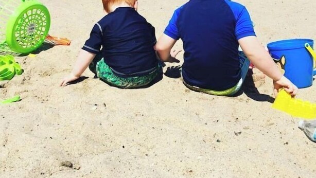 Zwei Kinder spielen an einem sandigen Strand, im Hintergrund sind Boote zu sehen.