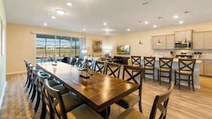 A spacious dining area with a long wooden table and multiple chairs, a kitchen with modern appliances, and a large window offering a view of the outdoors.