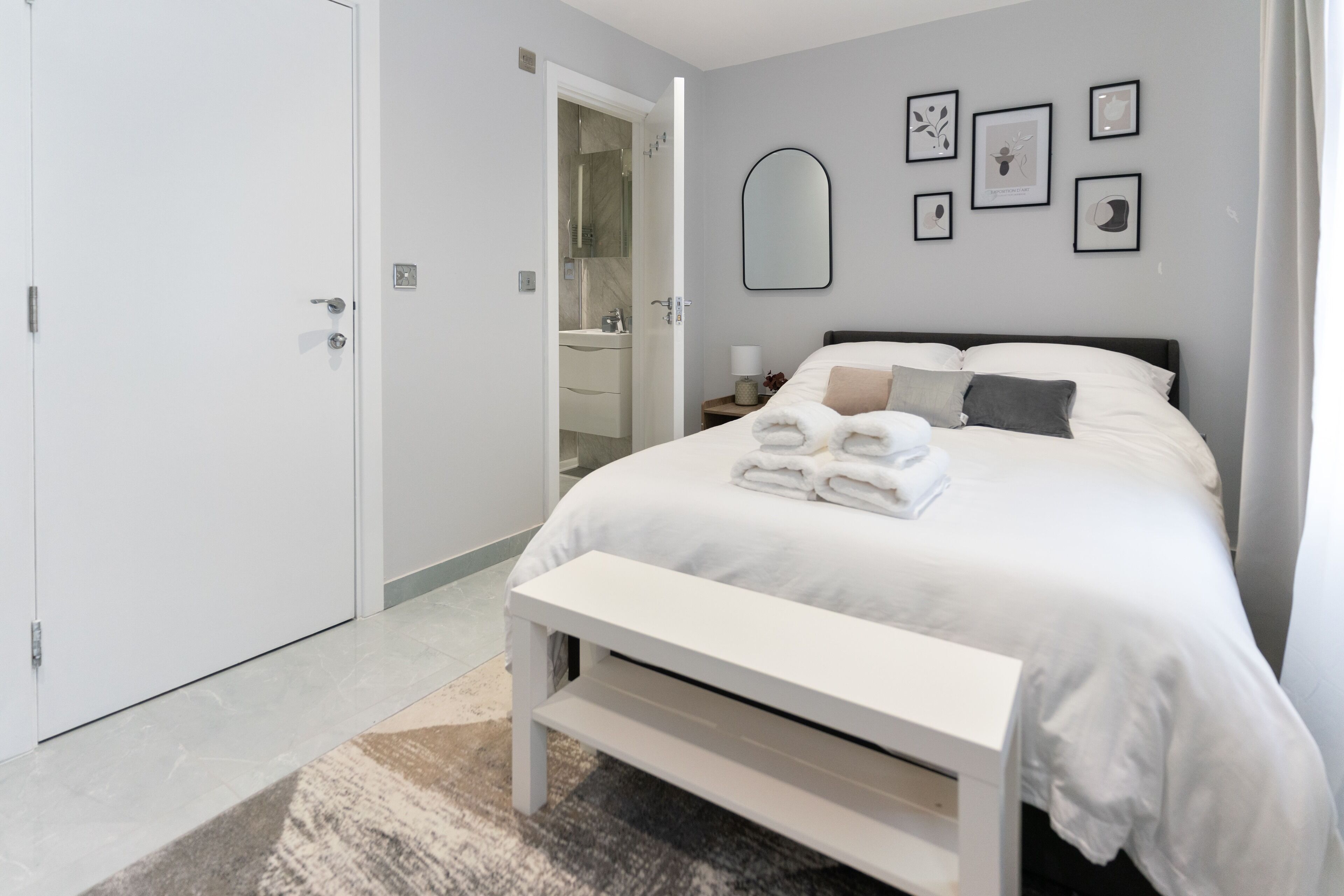 Un dormitorio moderno con una cama bien hecha, un banco y un baño visible a través de una puerta abierta.