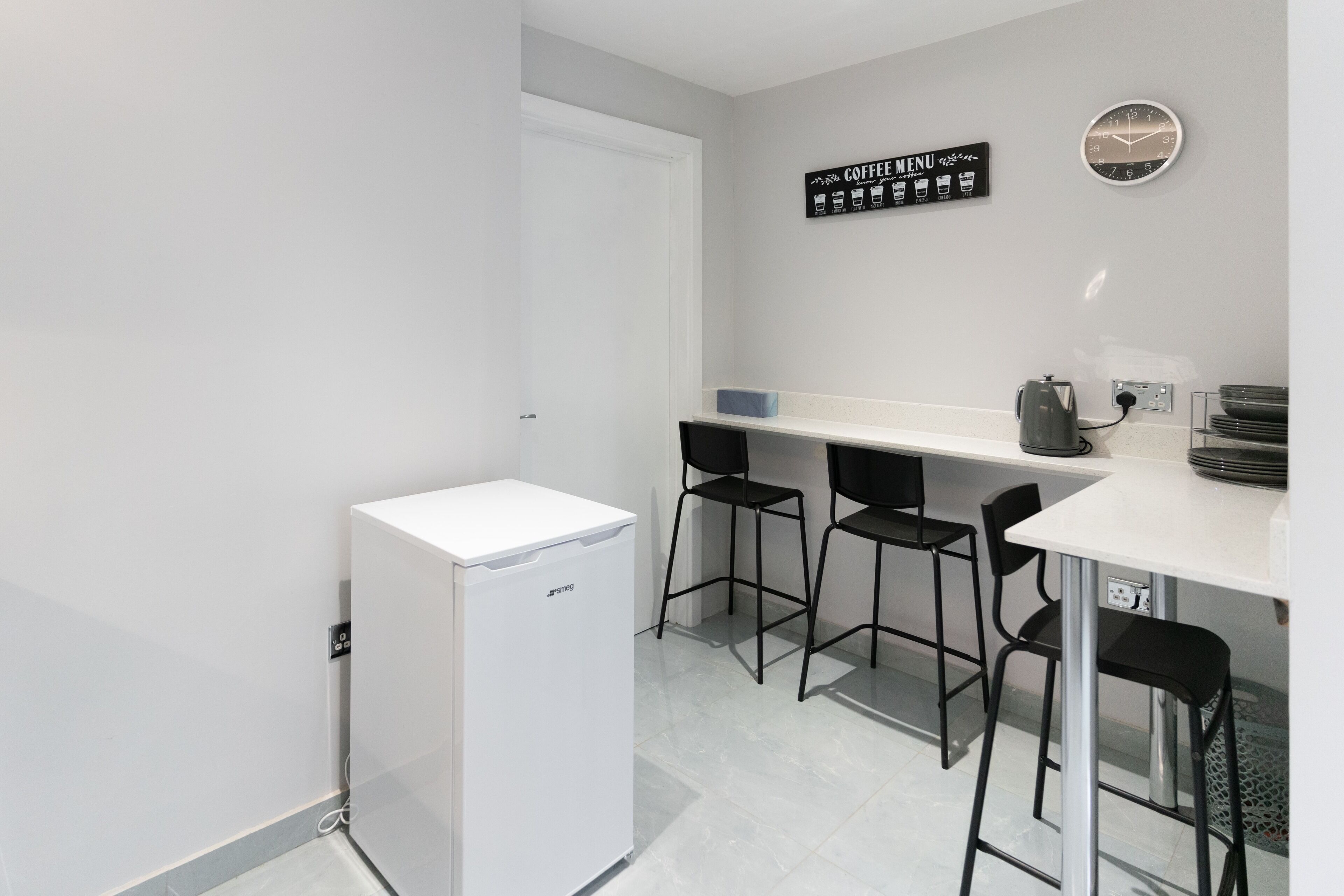 Une cuisine compacte avec un réfrigérateur blanc, un panneau indiquant le menu des cafés, une horloge et des tabourets de bar noirs.