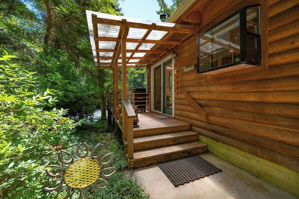 A wooden cabin with a covered porch, steps leading to a door, and a window with a view of the surrounding greenery. - McKenzie Bridge River's Edge Retreat, a cozy cabin on over two acres (McKenzie Bridge)