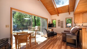 A modern living room with a wooden floor, a dining area with a table and chairs, a sofa, a television, and a kitchen area with wooden cabinets and a sink. - McKenzie Bridge River's Edge Retreat, a cozy cabin on over two acres (McKenzie Bridge)