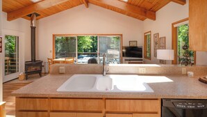 A kitchen with a large island, a wood-burning stove, and large windows overlooking a forested area. - McKenzie Bridge River's Edge Retreat, a cozy cabin on over two acres (McKenzie Bridge)