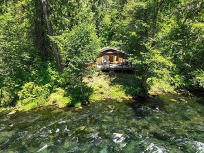 A cabin surrounded by dense forest, with a clear stream in front. - McKenzie Bridge River's Edge Retreat, a cozy cabin on over two acres (McKenzie Bridge)